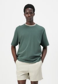Green short-sleeve t-shirt with a rounded neckline and white striped trim. The fabric appears soft, paired with beige shorts for a casual look.