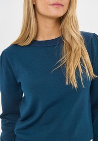 Szőke nő, aki kék kötött pulóvert visel, vállától a mellkasáig látható, egyszínű világos háttér előtt.