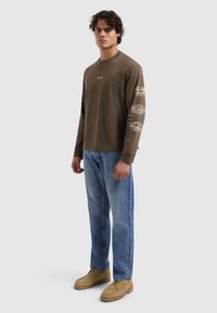 Olijfgroene shirt met lange mouwen en tekstgrafiek op de mouwen, gecombineerd met lichtblauwe spijkerbroek en beige schoenen. Eenvoudig ontwerp, katoenen materiaal.