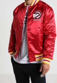 Červená saténová bomber bunda se žlutými a bílými proužkovanými žebrovanými manžetami a límcem. Na levé straně hrudníku je logo Atlanta Hawks.