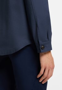 Tamsiai mėlyna ilgarankovė marškinėliai su lygiu audiniu, turintys užsegamą rankogalių sagtį ir šiek tiek lenktą apačią, derinami su atitinkančiomis tamsiomis kelnėmis.