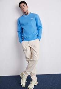 Blå sweatshirt med rund hals, logo på siden. Beige cargo bukser med lommer. Lysegrønne sneakers med et struktureret design. Blå baggrund.