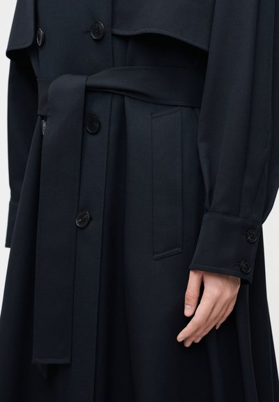 Gros plan d'une personne portant un trench-coat ceinturé bleu marine foncé avec des boutons, la main détendue le long du corps.