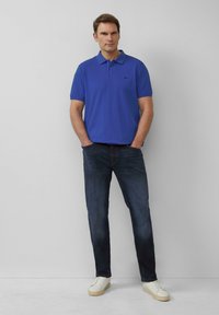 Blauwe polo shirt, korte mouwen, kraag, logo-detail. Donkern denim jeans, rechte pasvorm. Witte sneakers met een gestructureerde zool. Eenvoudig, strak design.