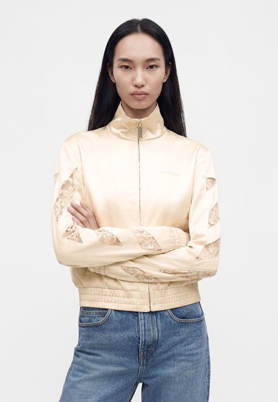 Jeune femme aux longs cheveux noirs, debout, les bras croisés, portant une veste en satin beige avec des découpes en dentelle et un jean bleu.