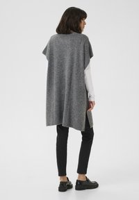 Harmaa neulottu poncho, jossa on löysä istuvuus, pyöreä pääntie ja sivuhalkiot, käytetään valkoisen pitkähihasen paidan ja mustien housujen päällä.
