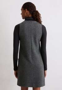 Grijs wolmix jurk met een getailleerde silhoeet, mouwloos ontwerp en achternaaddetail. G gedragen over een zwart langmouwtop.