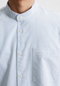 Camicia a righe blu e bianche con collo alla cinese, chiusura frontale con bottoni e taschino sinistro sul petto con testo ricamato.