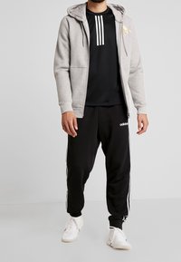 Sudadera gris con cremallera y bolsillos frontales sobre una camiseta negra con rayas blancas. Pantalones joggers negros con rayas laterales blancas y logo, combinados con zapatillas blancas.