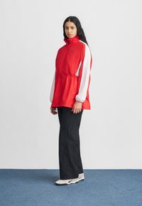 Jachetă pulover roșie cu mâneci albe, guler înalt și talie elastică. Completați ținuta cu pantaloni negri cu picioare largi și pantofi deschiși la culoare cu accente negre.