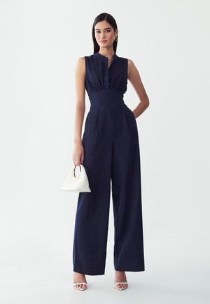 Femme portant une combinaison bleu marine sans manches et des talons blancs, tenant un petit sac à main blanc avec une poignée ronde dorée, debout devant un fond uni.