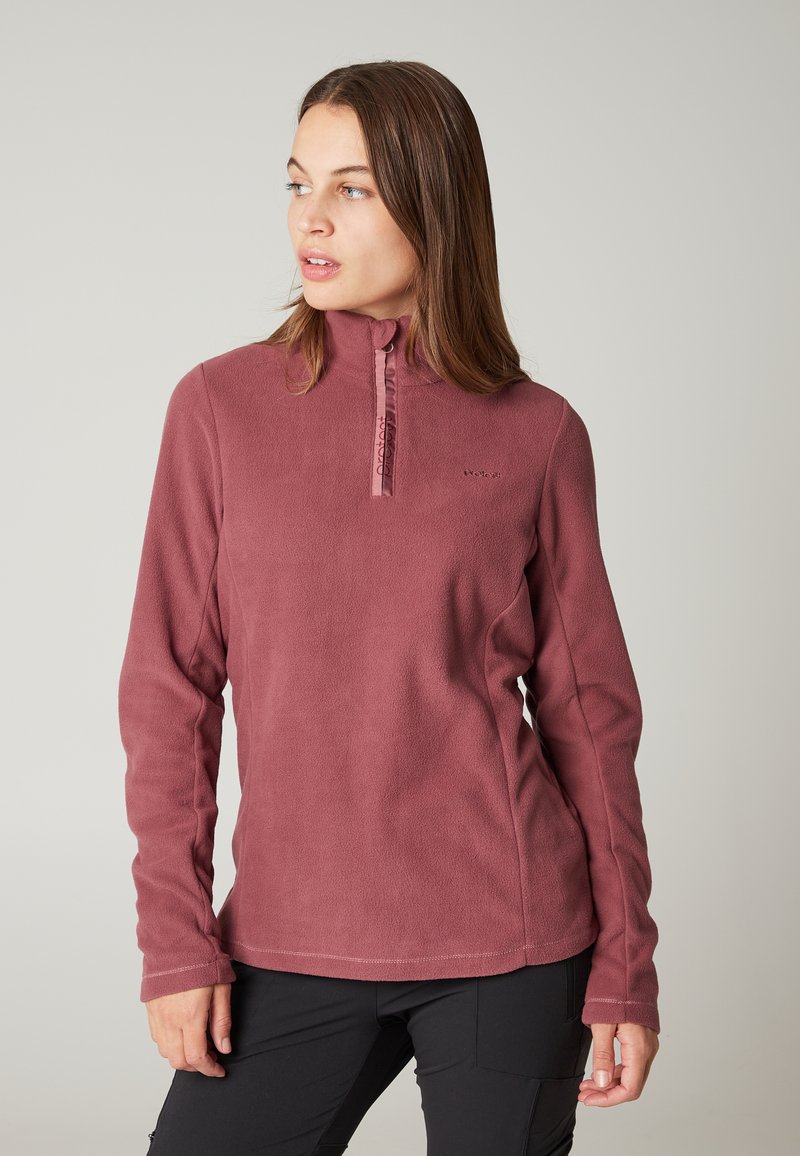 Kvinde iført en langærmet fleecepullover i støvet rosa med lynlås ved kraven, kigger til venstre, står mod en ensartet lysegrå baggrund.