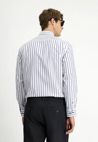 Camicia bianca con strisce verticali blu, maniche lunghe e colletto strutturato. Abbinata a pantaloni scuri. Indossati occhiali da sole neri.