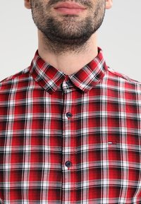 Bärtiger Mann trägt ein rot-schwarz-weißes kariertes Hemd mit Knöpfen, dessen Kragen geschlossen ist.