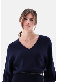 Marineblauer Strickpullover mit tiefem V-Ausschnitt, langen Ärmeln, strukturierter Stoff und tailliertem Bund, betont durch einen schmalen Gürtel.