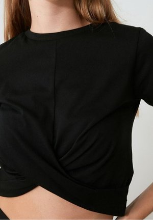 Kobieta nosząca czarny krótki crop top z przekręconym detalem z przodu i okrągłym dekoltem.