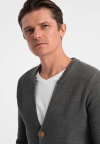 Grå stickad cardigan med V-ringning och en träknapp. Buren över en vit T-shirt med V-ringning. Slät textur, avslappnad passform.