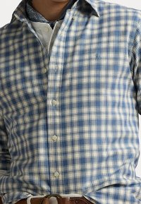 Sininen ja valkoinen ruudullinen napitettava paita, jossa on kaulus, pitkät hihat ja pieni logo yksityiskohtana rinnassa. Kangas näyttää olevan teksturoitu.