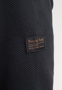 Tessuto blu scuro con un motivo a spina di pesce testurizzato. Un'etichetta rettangolare riporta la dicitura "Petrol Ind" con dettagli sulle sue origini in denim.