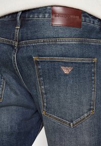Tmavě modré džíny z denimu s hnědou koženou etiketou s nápisem "EMPORIO ARMANI." Výrazné růžové logo na zadní kapse a žluté prošívání.