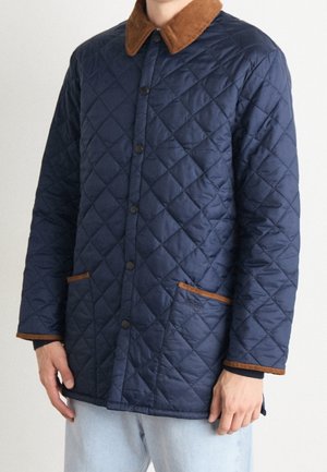Homme portant une veste matelassée bleu marine avec un col en velours côtelé brun et une finition de poche, debout les mains détendues sur les côtés.