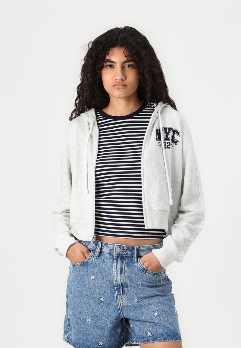 Felpa grigia con cappuccio e zip frontale, con coulisse e stampa "NYC". Abbinata a un crop top a righe blu navy e bianche e a pantaloni di jeans ricamati.