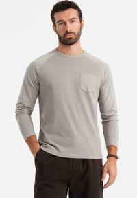 Harmaa pitkähihaisen T-paidan, jossa on teksturoitu kangas, ribattu muotoilu ja rintatasku. Korostettuna raglan-hihoilla ja pyöreällä kaula-aukolla.