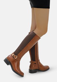 Botas marrones de cuero hasta la rodilla con un panel de tela marrón texturizado, una cremallera lateral y un pequeño detalle de hebilla en el tobillo.