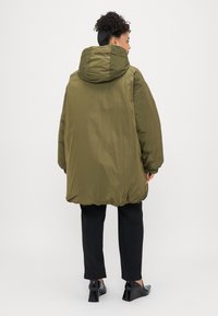 Alyvuogių spalvos pūkinis švarkas su gobtuvu, turintis laisvą siluetą ir tekstūruotą apdailą. Derinamas su juodomis prigludusiomis kelnių ir aukštakulniais bateliais.