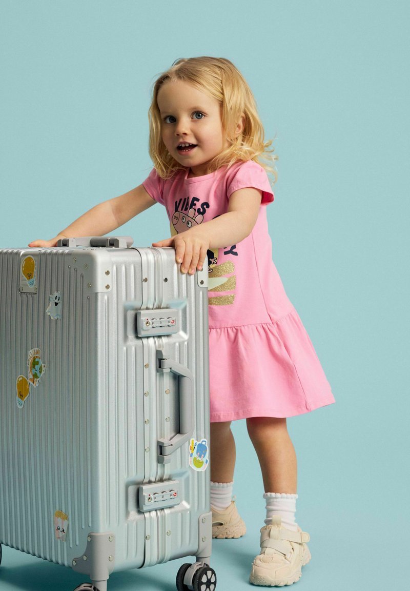 Jeune fille en robe rose debout à côté d'une valise argentée décorée d'autocollants de voyage colorés sur un fond bleu clair.