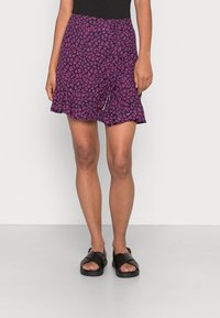 Jupe à imprimé floral en violet foncé avec des fleurs roses, ourlet froncé et taille ajustable. Associée à des sandales noires.