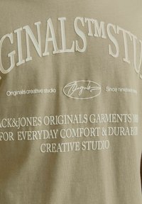 Lichtbruine katoenen t-shirt met witte geborduurde tekst. Bevat de merknaam, slogan en logo in verschillende lettergroottes. Eenvoudig ontwerp, casual pasvorm.