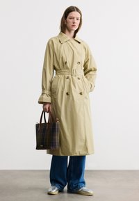 Femme portant un trench-coat beige, jean large bleu, baskets argentées, tenant un sac à main en tissu à carreaux foncé avec des poignées marron, debout sur un sol gris.