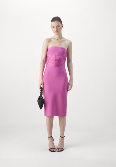 Fitted pink strapless dress with gathered texture at the bodice and a knee-length hem. Accessorized with a small black handbag.