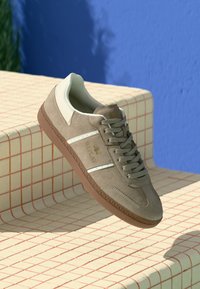 Tenisi din suede bej cu talpă brună, dungi albe și logo-ul "Replay", așezați pe o scară din faianță crem cu un fundal de perete albastru.
