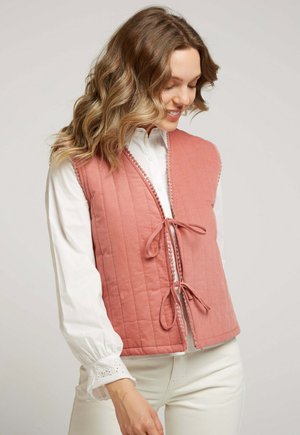 Gilet rose matelassé avec une fermeture à l'avant nouée, présentant un tissu texturé et une bordure décorative sur les bords. Porté par-dessus une chemise blanche.