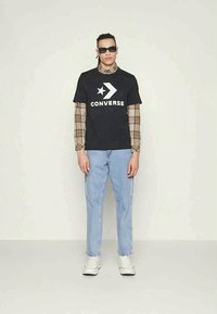 Black cotton t-shirt with white "CONVERSE" logo and star design, layered over a checked long-sleeve shirt, paired with light blue jeans and white sneakers.