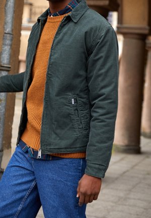 Person iført blå jeans, grøn jakke og orange sweater, står ude ved stenrammer og holder en metalstang.