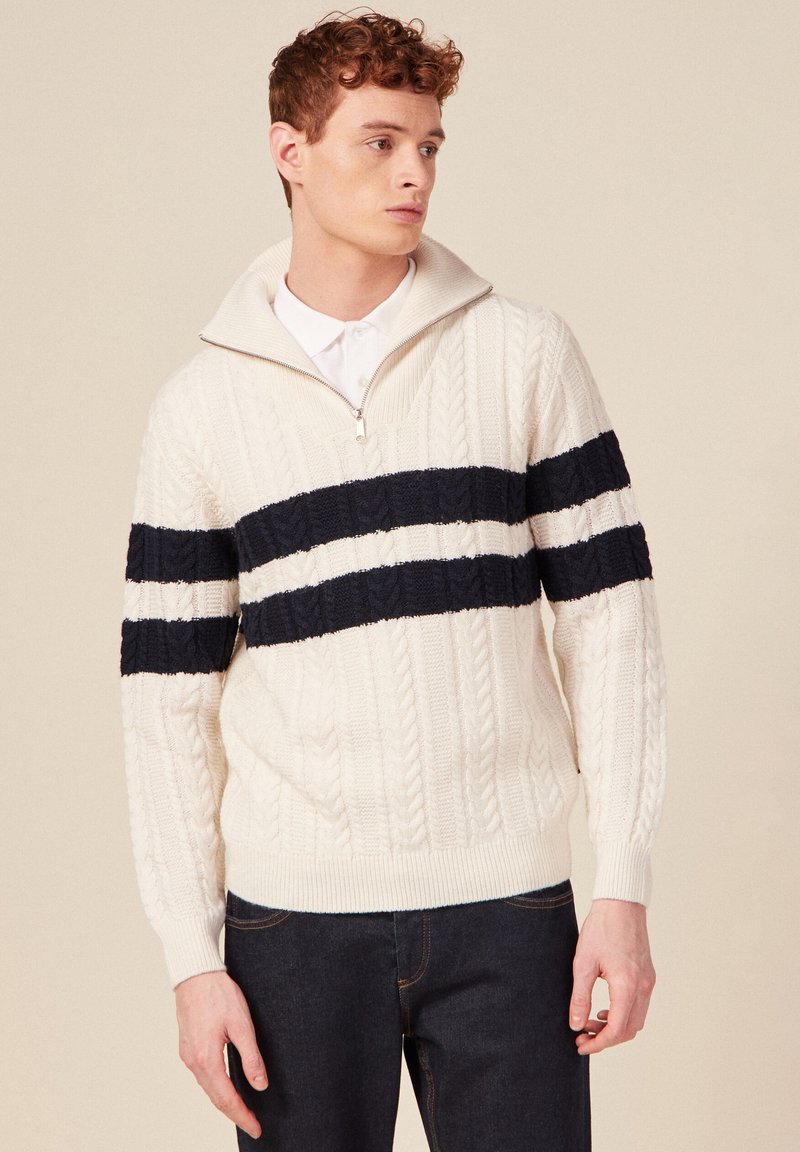 Kabelstrickpullover aus cremiger Wolle mit marineblauen Streifenakzenten. Verfügt über einen mit Reißverschluss versehenen hohen Kragen und gerippte Bündchen, die strukturierte Details bieten.