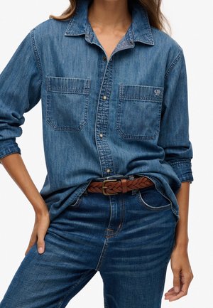 Woman wearing blue denim button-up shirt with two chest pockets, paired with blue jeans and a brown braided belt, posing with hands on hips.