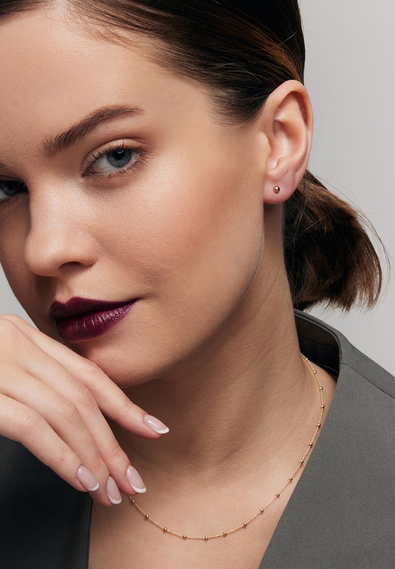 Złote kolczyki wkrętki, delikatny naszyjnik z złotych koralików, strój w neutralnych odcieniach, gładka tekstura skóry oraz ciemna burgundowa pomadka podkreślają rysy twarzy.