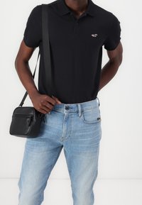 Polo negro de manga corta con un pequeño logo, jeans de mezclilla de color azul claro y un bolso bandolera de cuero negro.