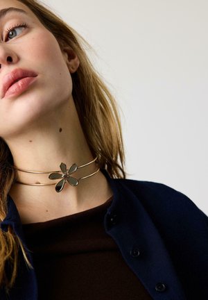 Giovane donna che indossa un choker doppio in oro con un pendente a specchio a forma di fiore, vestita con una maglia scura e un cardigan blu navy con bottoni.