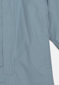 Giacca impermeabile azzurro chiaro con una texture liscia, dotata di una tasca sul petto e cuciture a contrasto lungo le cuciture.