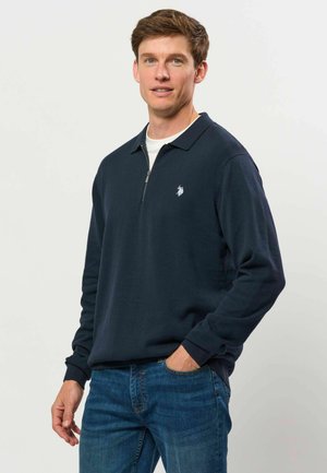 Young man with short brown hair wearing a navy blue long-sleeve polo shirt and blue jeans standing with one hand in pocket.