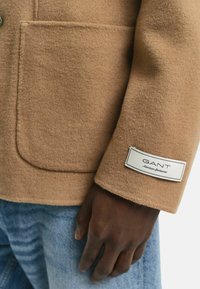 Bruine wollen jas met een gestructureerde afwerking, voorzien van een opgestikte zak en een label op de manchette met de tekst "GANT American Sportswear."