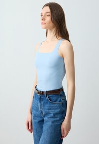 Top aderente azzurro chiaro con scollatura quadrata, abbinato a jeans blu a vita alta e una cintura marrone. Texture liscia e design essenziale.