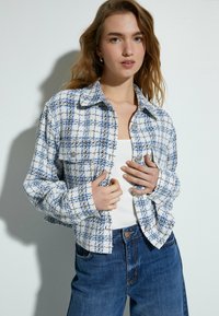 Blauw-wit geruite cropped jas met een kraag, twee voorzakken en knoopsluitingen, gecombineerd met een geribbelde witte top en blauwe jeans.