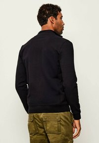 Felpa nera con colletto e polsini a coste, caratterizzata da una texture liscia e una vestibilità comoda. Indossata con pantaloni cargo verde oliva.