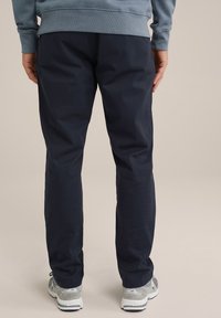 Jūras zila ikdienas bikses taisnā griezumā, ar gludu tekstūru, sānu kabatām un jostasvietu, valkātas ar pelēkiem sporta apaviem.